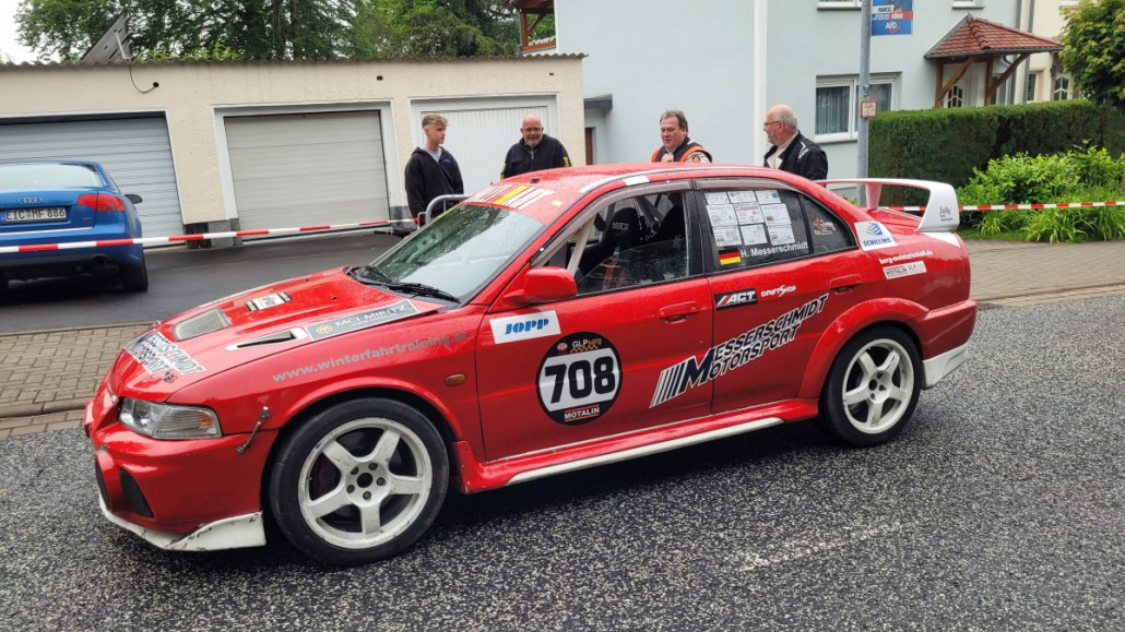 Auf einer Straße parkt ein roter Rennwagen mit der Nummer 708 auf der Tür. In der Nähe stehen mehrere Personen, im Hintergrund sind Wohnhäuser und Garagen zu sehen.