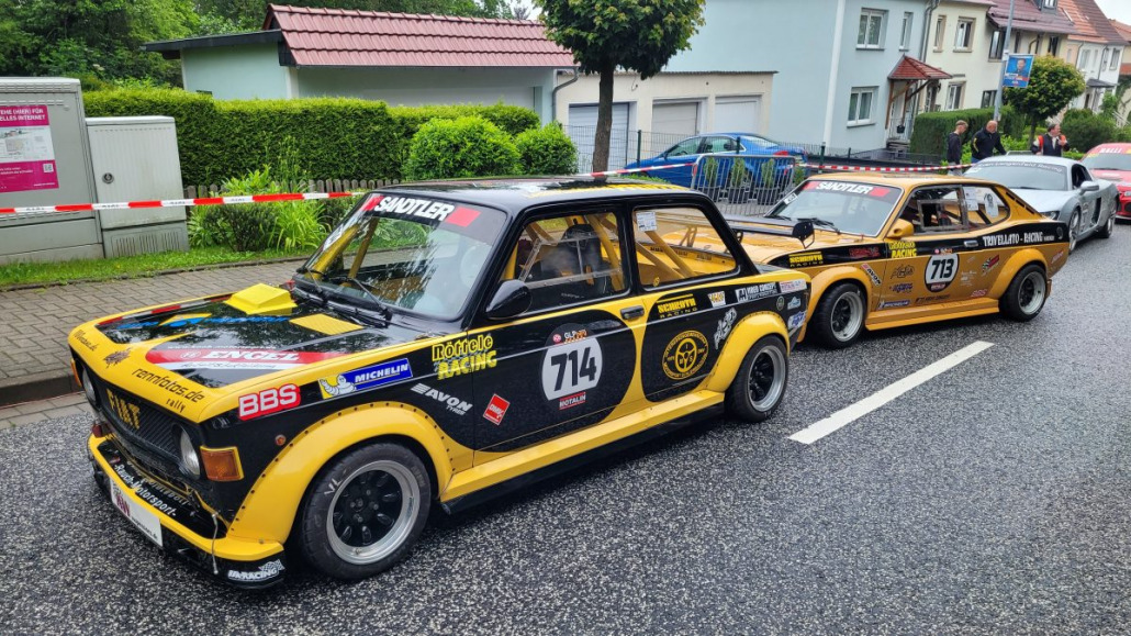 Zwei schwarz-gelbe Rennwagen stehen auf einer Straße mit Häusern im Hintergrund. Die Autos tragen verschiedene Sponsorenlogos und Startnummern, vorne 714 und hinten 713.