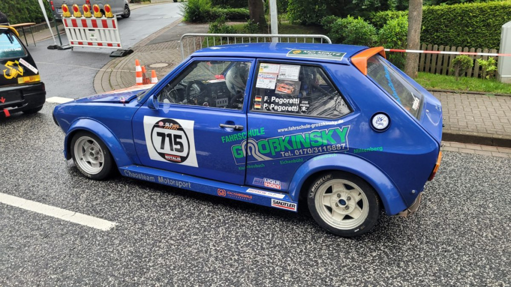 Blaues Rallyeauto mit der Nummer 715, gesponsert von Godrinksky und anderen Marken, geparkt auf einer asphaltierten Straße mit Absperrungen und üppigem Grün im Hintergrund.