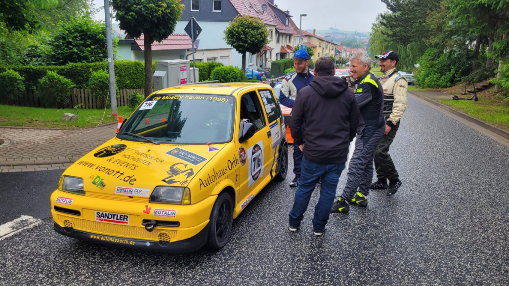 Eine Gruppe von Menschen steht neben einem gelben Rallyeauto, das in einer Wohnstraße mit Häusern und Bäumen im Hintergrund geparkt ist.