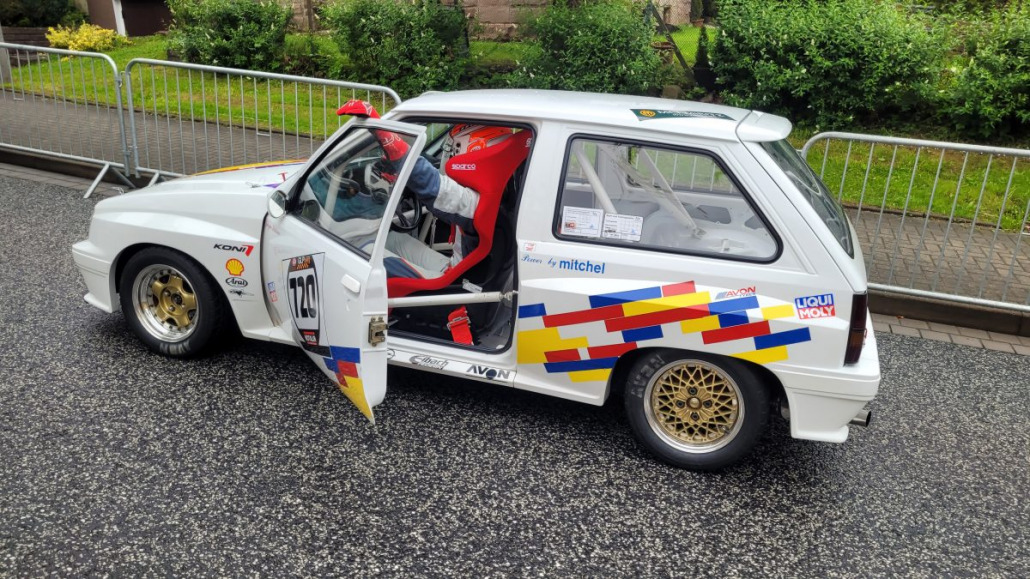 Ein weißes Rallyeauto mit bunten Aufklebern und der Nummer 243 sowie offener Fahrertür ist auf einer asphaltierten Straße neben einer Metallabsperrung geparkt, im Hintergrund sieht man Grünflächen.