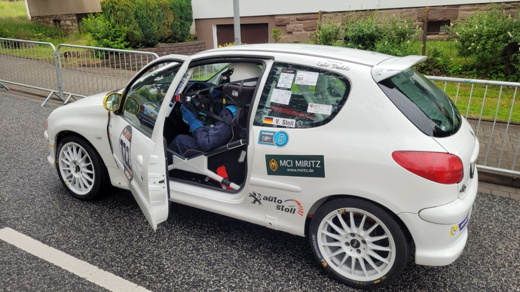 Auf einer nassen Straße geparktes weißes Rallyeauto mit geöffneter Fahrertür. Auf der hinteren Seitenscheibe und der hinteren Stoßstange sind Aufkleber von Sponsoren und Nummernschilder angebracht.