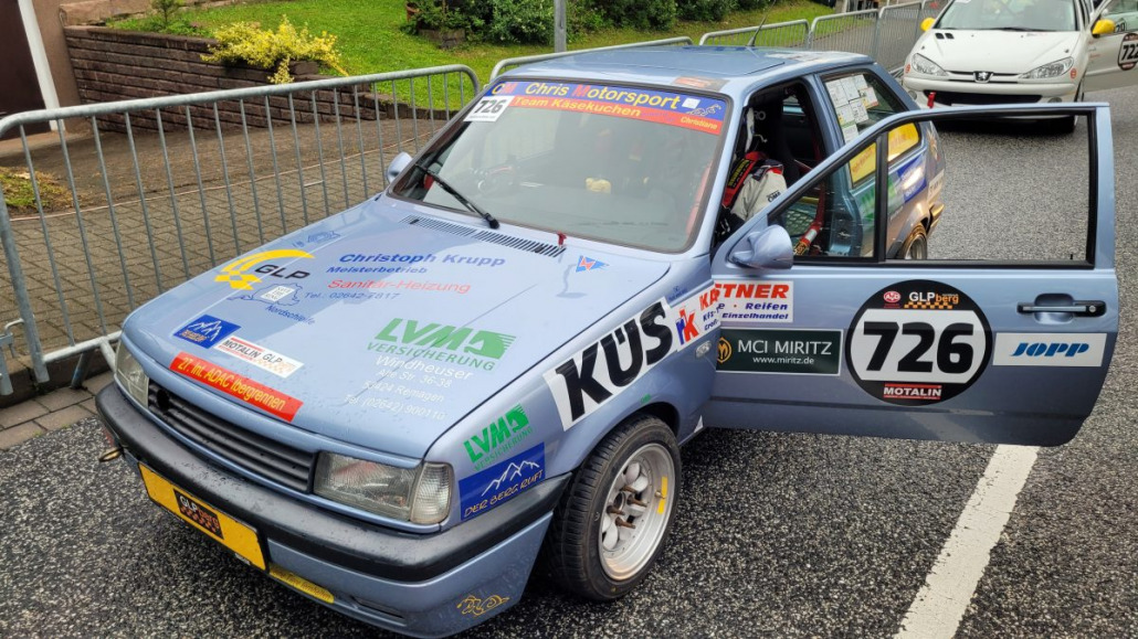 Ein hellblaues Rallyeauto mit der Seitennummer 726 und verschiedenen Sponsorenlogos parkt auf einer Straße, die Beifahrertür ist geöffnet.