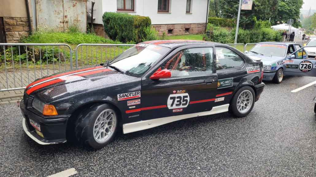 Zwei Rennwagen stehen auf einer nassen Straße. Das nächste Auto mit der Nummer 735 ist schwarz mit roten und orangen Streifen und mehreren Sponsorenlogos. Das zweite Auto mit der Nummer 726 ist teilweise hinter dem ersten zu sehen.