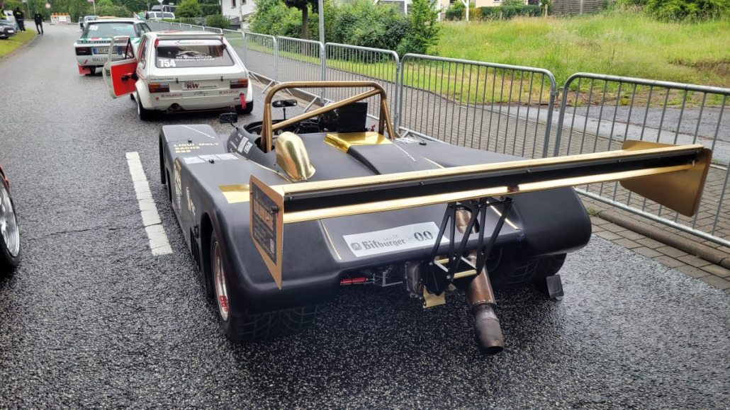 Mehrere historische Rennwagen, darunter ein schwarzer Roadster mit großem Heckflügel, stehen in einer Reihe auf einer nassen Straße, getrennt durch Metallbarrieren.