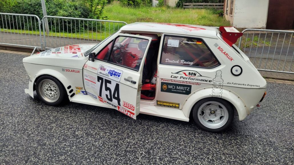 Ein weißes Rallyeauto mit der Nummer 754 auf der Tür, geparkt auf einer nassen Straße. Das Fahrzeug ist mit Sponsorenaufklebern bedeckt und die Fahrertür ist offen. Im Hintergrund sind Metallbarrikaden und eine Rasenfläche zu sehen.