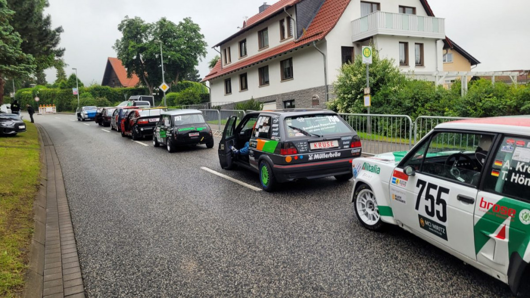 Mehrere Rennwagen stehen in einer Wohnstraße aufgereiht und sind in verschiedenen bunten Designs gestaltet. Die Umgebung ist von Häusern, Grünflächen und einem bewölkten Himmel geprägt.