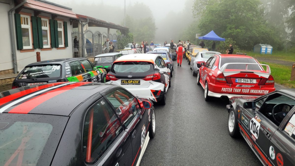 Eine Gruppe von Rennwagen steht auf einer nassen Straße in der Nähe eines Gebäudes mit gewölbten Fenstern, umgeben von Nebel. Menschen in Rennanzügen stehen um die Fahrzeuge herum.