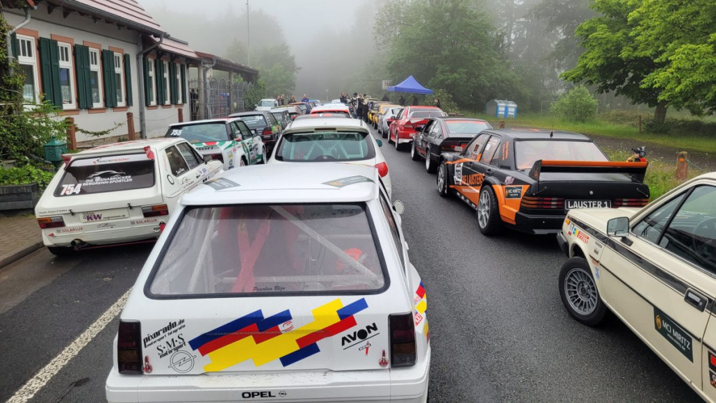 Eine Reihe von Rallyeautos parkt auf einer nebligen Straße in der Nähe eines Gebäudes. Die Fahrzeuge sind mit verschiedenen bunten Aufklebern versehen und im Hintergrund stehen Menschen unter einem blauen Baldachin.