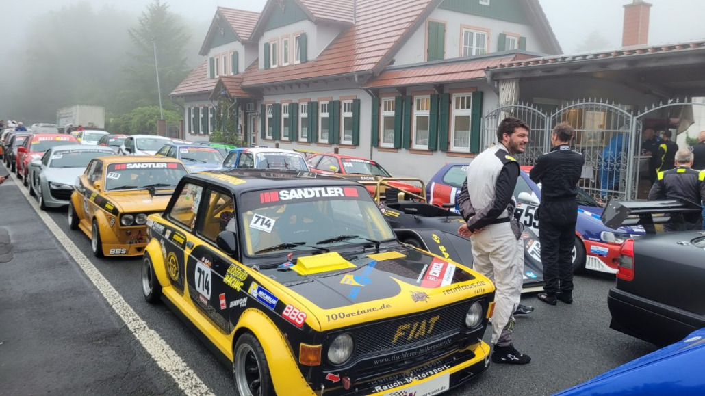 Eine Reihe farbenfroher Oldtimer-Rennwagen und Fahrer in Rennanzügen an einem nebligen Tag, geparkt neben einem traditionellen Gebäude mit Ziegeldach. Auf einem gelben Auto im Vordergrund ist die Nummer 714 zu sehen.