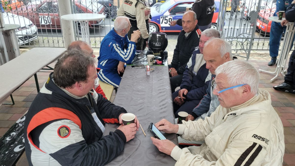 Eine Gruppe Männer in Rennanzügen sitzt an einem Tisch im Freien und unterhält sich. Einige halten Kaffeetassen in den Händen, einer benutzt ein Tablet. Im Hintergrund sind Rennautos zu sehen.