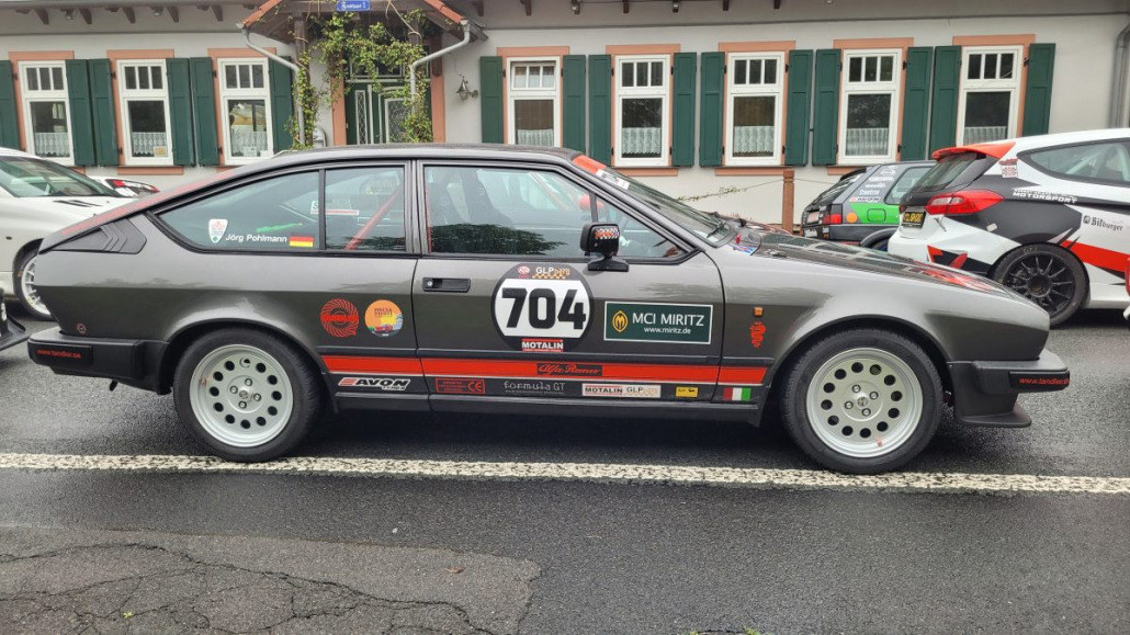 Ein geparkter, grauer Oldtimer-Sportwagen mit der Nummer 704 und verschiedenen Sponsorenaufklebern an der Seite. Im Hintergrund ist teilweise ein weiteres Auto zu sehen.