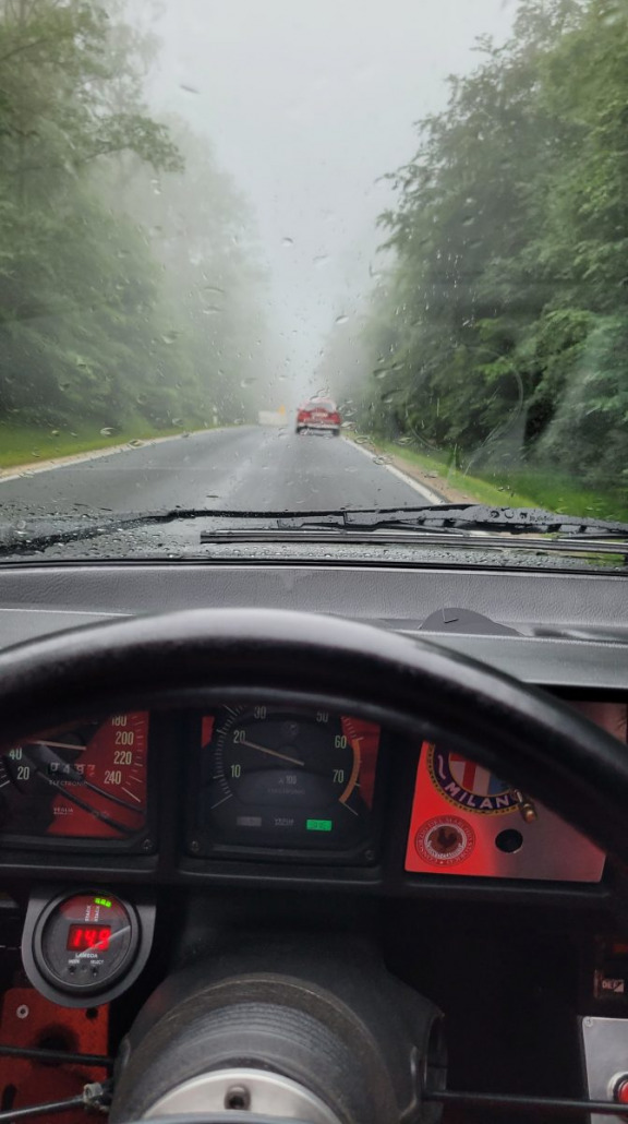 Blick durch die Windschutzscheibe eines Autos an einem regnerischen Tag. Die Windschutzscheibe ist nass von Regentropfen und die Straße vor uns ist neblig. Der Tacho zeigt 45 km/h an und in der Ferne ist ein rotes Auto zu sehen.