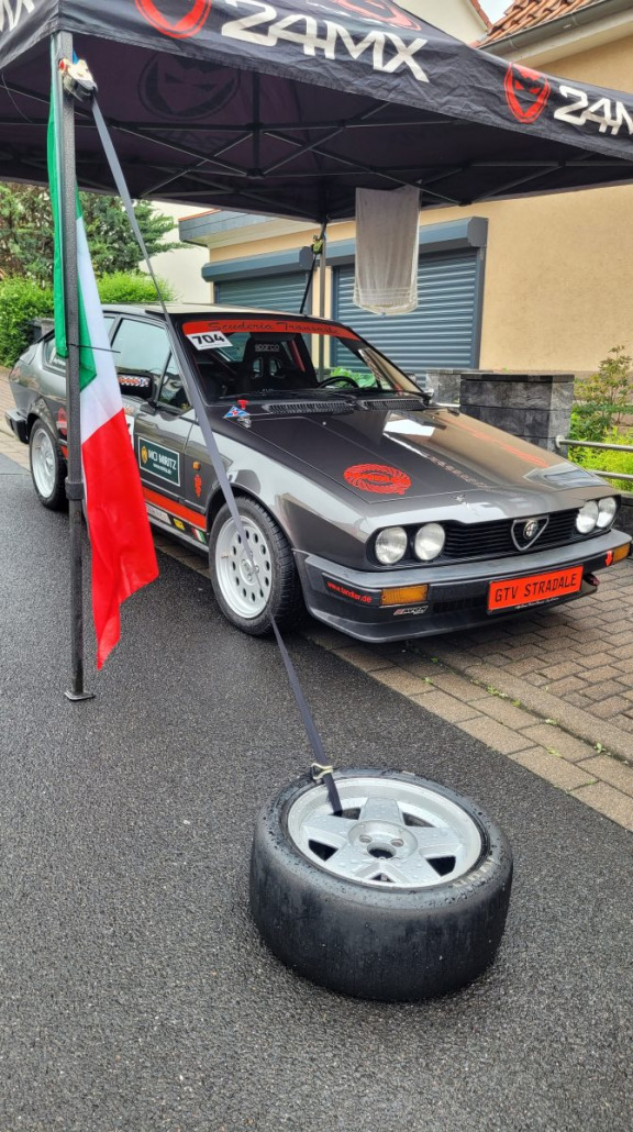 Ein grauer Alfa Romeo GTV Stradale-Oldtimer, geparkt unter einem schwarzen Z4MX-Überdach mit Rennaufklebern, neben einer italienischen Flagge und einem auf dem Boden platzierten Reifen.