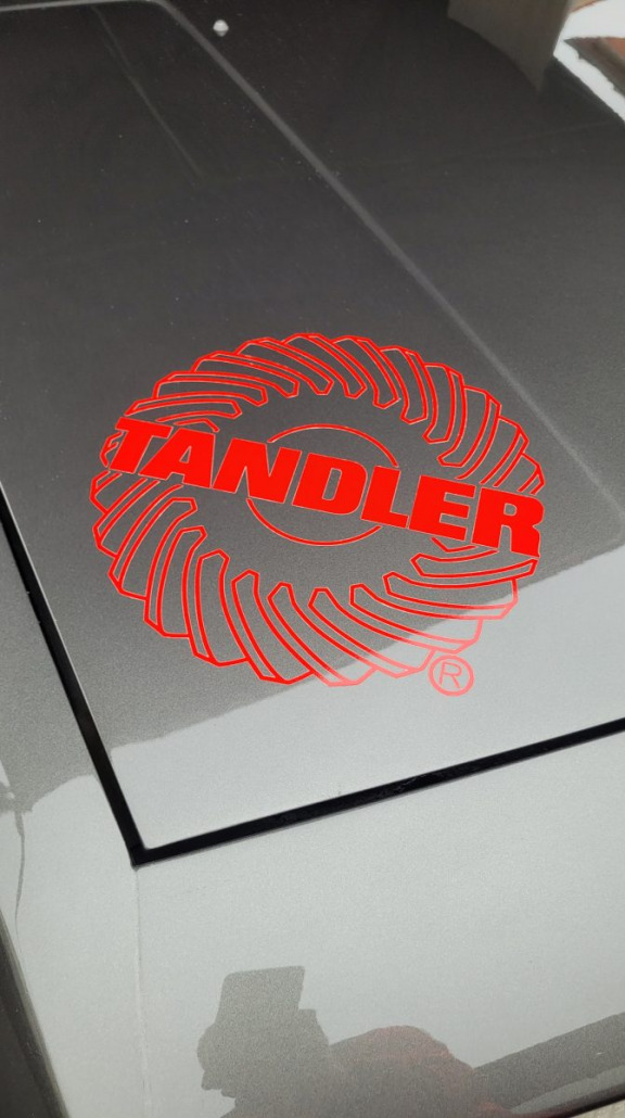 Nahaufnahme einer Motorhaube eines Autos mit einem roten, runden Logo und dem Wort „TANDLER“ in fetten Buchstaben.