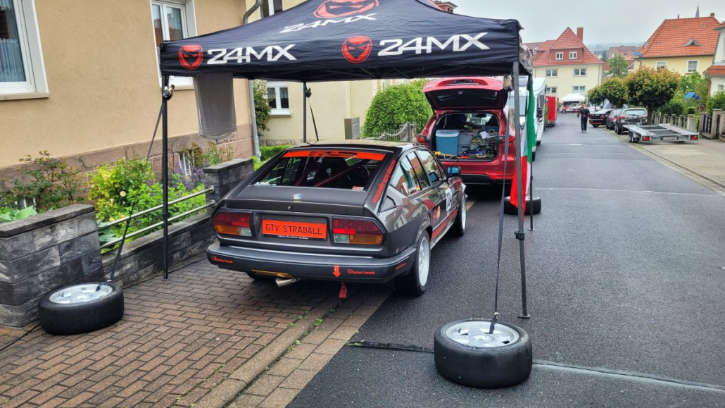 Ein grauer Alfa Romeo GTV Stradale ist mit geöffnetem Kofferraum unter einem 24MX-Überdach geparkt. Ersatzreifen liegen neben den Stützpfosten des Überdachs in einer Wohnstraße.