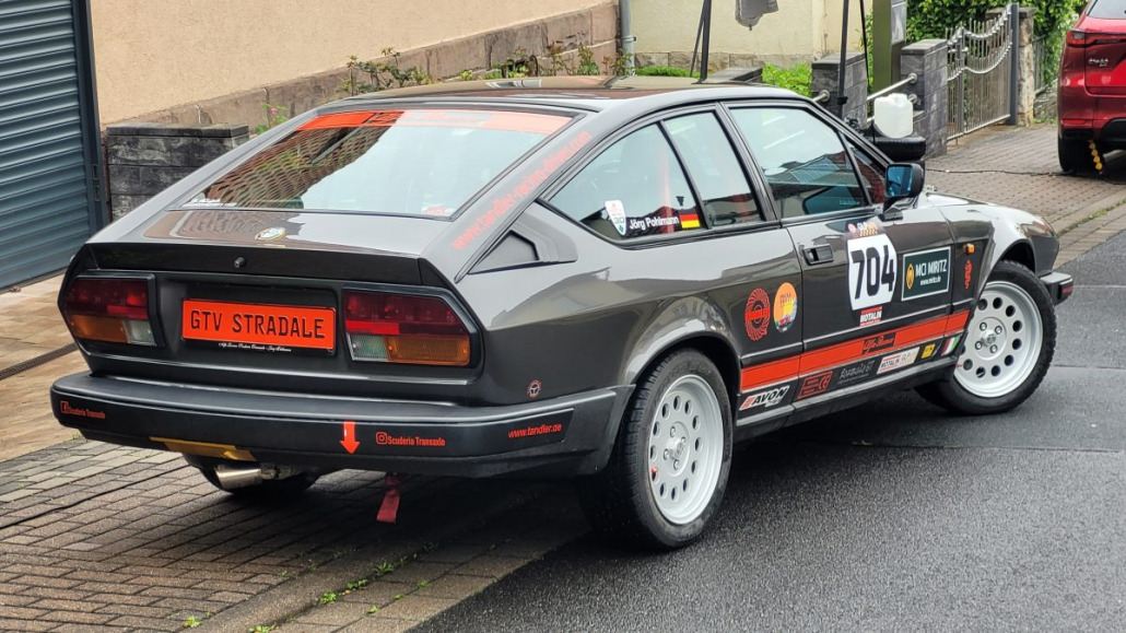 Ein gereinigtes und poliertes dunkelgraues Rallyeauto Alfa Romeo Alfasud GTV Stradale mit dem Aufkleber mit der Nummer 704 und verschiedenen Sponsorenaufklebern, geparkt auf einer nassen Straße.