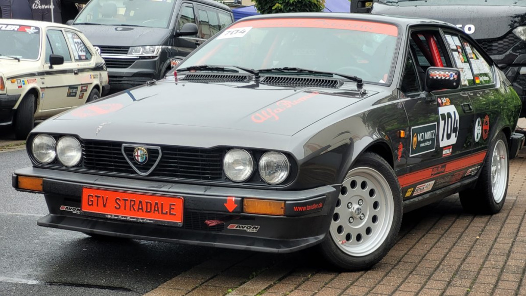 Ein schwarzer Alfa Romeo GTV Stradale mit Rennaufklebern steht neben anderen Autos auf einer Straße. Das Auto hat die Nummer 704 auf der Windschutzscheibe und einen markanten Kühlergrill mit runden Scheinwerfern.