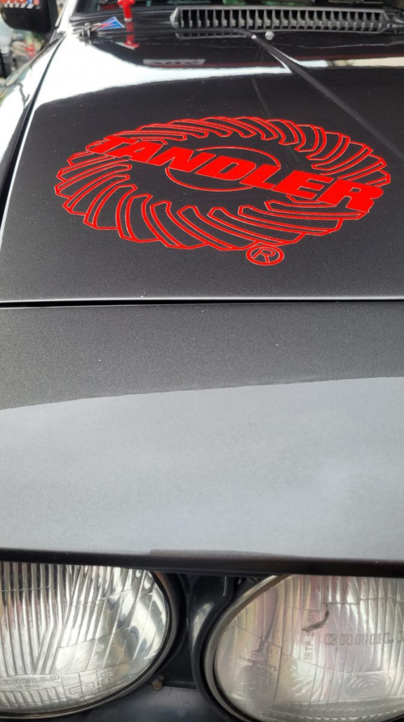 Nahaufnahme einer Motorhaube mit einem rot-schwarzen Emblem, auf dem das Wort „ENOLER“ steht, umgeben von wirbelnden Grafiken. Darunter sind zwei Scheinwerferlampen teilweise sichtbar.