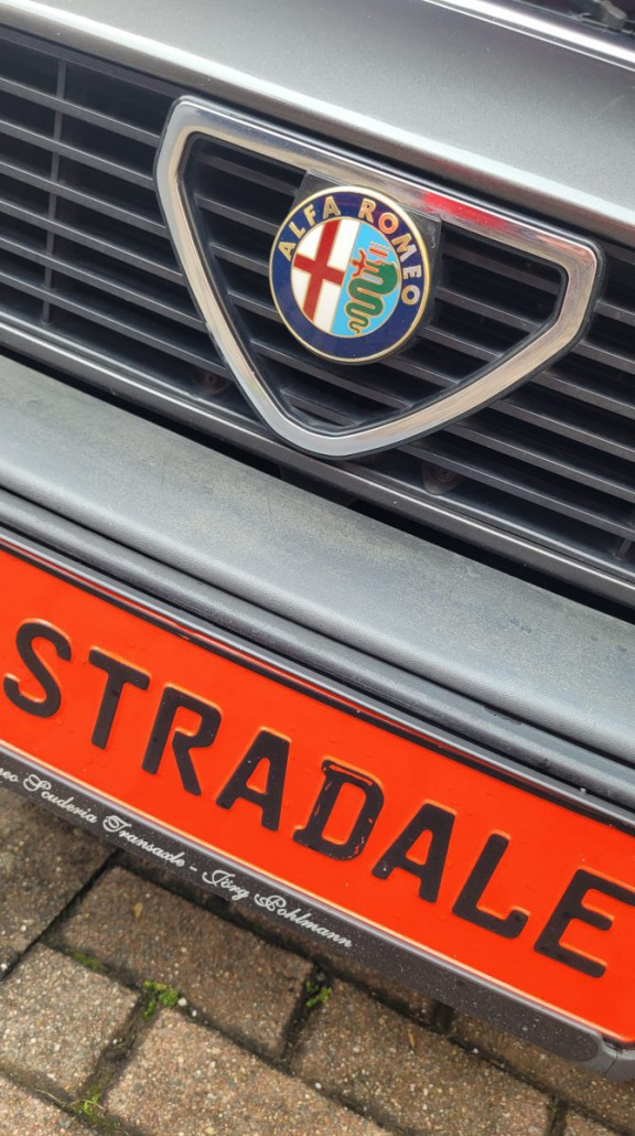 Nahaufnahme des Kühlergrills eines Alfa Romeo mit dem Alfa Romeo-Emblem und einem roten Nummernschild mit dem Wort „STRADALE“. Das Auto ist auf einem Kopfsteinpflaster geparkt.