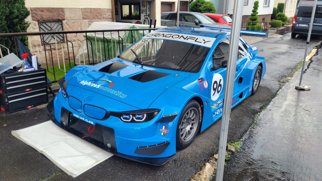 Ein blaues Rennauto mit der Nummer 96 auf den Türen steht im Freien auf einem Bürgersteig, teilweise geschützt durch ein weißes Vordach. Die Fronthaube ist mit mehreren Lufteinlässen ausgestattet und Sponsorenaufkleber sind sichtbar.