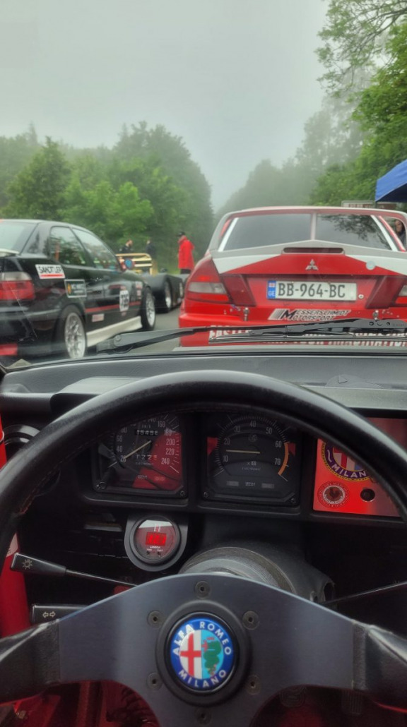 Blick aus dem Inneren eines klassischen Alfa Romeo-Rennwagens auf eine Reihe von Autos vor uns an einem nebligen Tag, darunter ein roter Mitsubishi Lancer mit dem Nummernschild BB-964-BC.