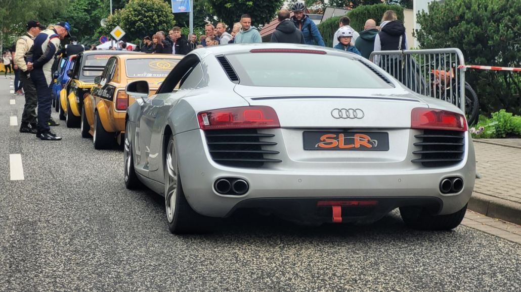 Ein silberner Audi-Sportwagen mit dem Kennzeichen „SLR“ steht neben zwei gelben Fahrzeugen auf einer Straße. In der Nähe hat sich eine Menschenmenge versammelt, einige stehen, andere unterhalten sich.