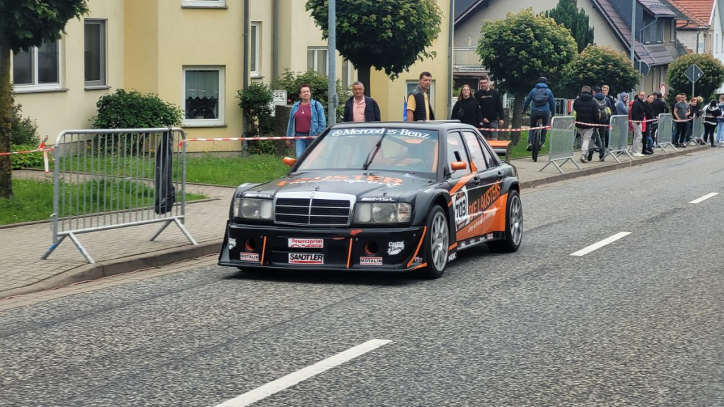 Ein schwarz-orangefarbener Mercedes-Benz-Rennwagen parkt auf einer Straße. Auf beiden Seiten der Straße stehen Zuschauer hinter Absperrungen.