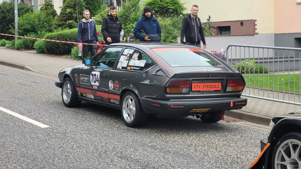 Ein grauer GTV Stradale mit der Startnummer 104 steht auf einer Straße. Die hinteren Seitenfenster sind mit mehreren Aufklebern bedeckt. In der Nähe stehen vier Männer auf dem Bürgersteig, zwei von ihnen lächeln.