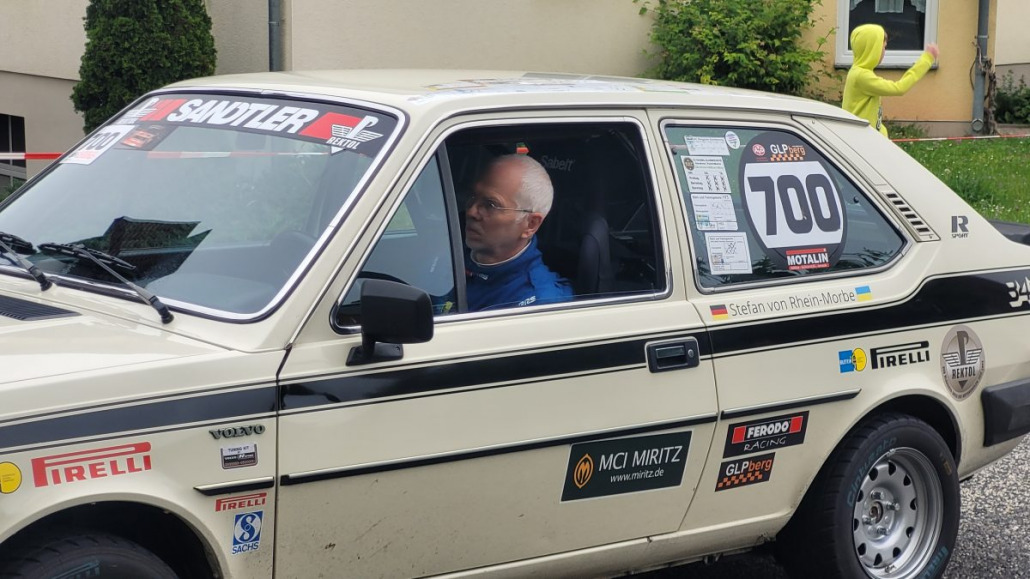 Ein Mann sitzt in einem Oldtimer-Rallyeauto mit der Nummer 700 an der Seite und bereitet sich auf ein Rennen vor. Das Auto trägt mehrere Sponsorenlogos, darunter Pirelli und MCI Miritz. Im Hintergrund ist eine Person in einem gelben Anzug zu sehen.