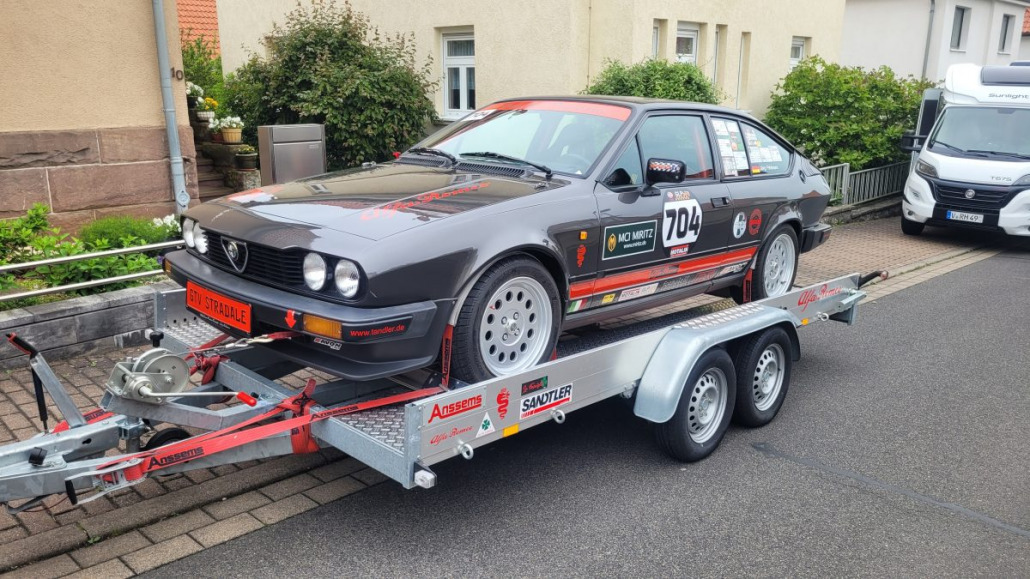 Ein schwarz-roter Rennwagen mit der Nummer 704 steht festgemacht auf einem Anhänger in einem Wohngebiet, neben einem Wohnmobil.