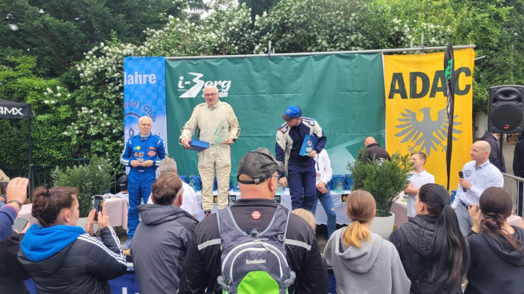 Drei Rennfahrer nehmen auf einem Podium ihre Auszeichnungen entgegen, umringt von Zuschauern und Fotografen. Im Hintergrund hängen Banner mit den Aufschriften "L-Zberg" und "ADAC". Die Veranstaltung findet offenbar im Freien statt.