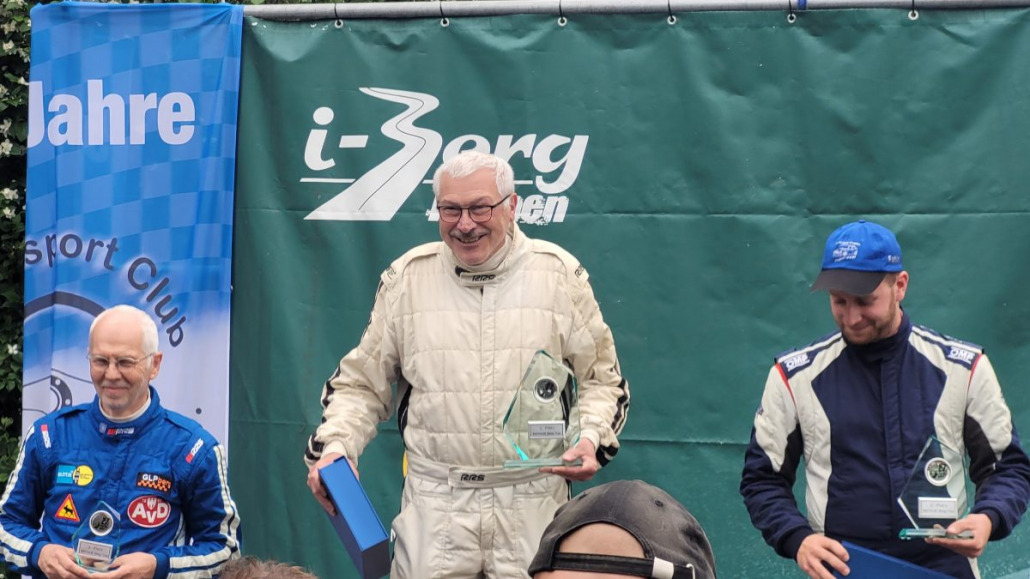 Drei Männer stehen mit Trophäen auf einem Podium und feiern ein Motorsportereignis. Der Mann in der Mitte lächelt und hält eine größere Trophäe, während die Männer auf beiden Seiten kleinere Trophäen halten.