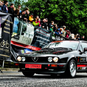 Ein schwarzer Alfa Romeo GTV Stradale mit Rennaufklebern fährt während einer Rennveranstaltung an einer Menge Zuschauer vorbei. Im Hintergrund sind Banner und Bäume zu sehen.