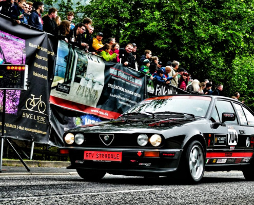 Ein schwarzer Alfa Romeo GTV Stradale mit Rennaufklebern fährt während einer Rennveranstaltung an einer Menge Zuschauer vorbei. Im Hintergrund sind Banner und Bäume zu sehen.