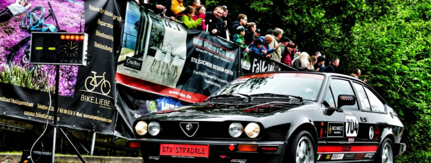 Ein schwarzer Alfa Romeo GTV Stradale mit Rennaufklebern fährt während einer Rennveranstaltung an einer Menge Zuschauer vorbei. Im Hintergrund sind Banner und Bäume zu sehen.