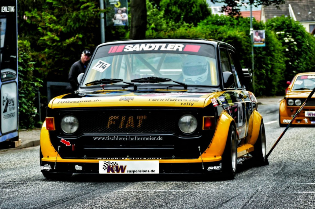 Ein gelb-schwarzes Rennauto mit Fiat-Logo, Nummer 714 und mehreren Sponsorenaufklebern steht auf einer Straße. Im Inneren sitzt ein Fahrer mit Helm. Im Hintergrund ist ein weiteres Auto zu sehen.