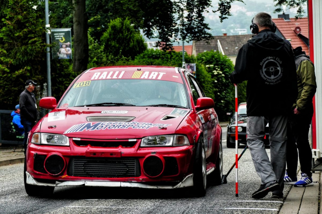 Ein rotes Rallyeauto mit der Aufschrift „RALLIART“ auf der Windschutzscheibe steht auf einer Straße, umgeben von Menschen, von denen einer ein Headset trägt und einen Messstab hält. Im Hintergrund sind Bäume und Häuser zu sehen.