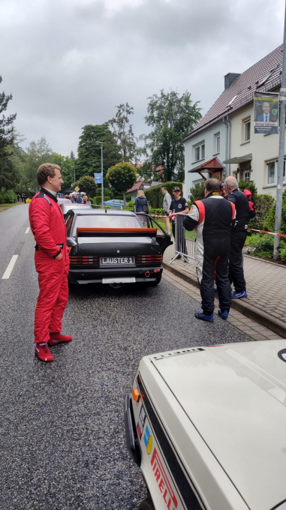 Ein Mann im roten Rennanzug steht neben einem Oldtimer auf einer Straße, während sich zwei andere in schwarzen Anzügen neben dem Auto unterhalten. Im Hintergrund sind weitere Zuschauer und Häuser zu sehen.