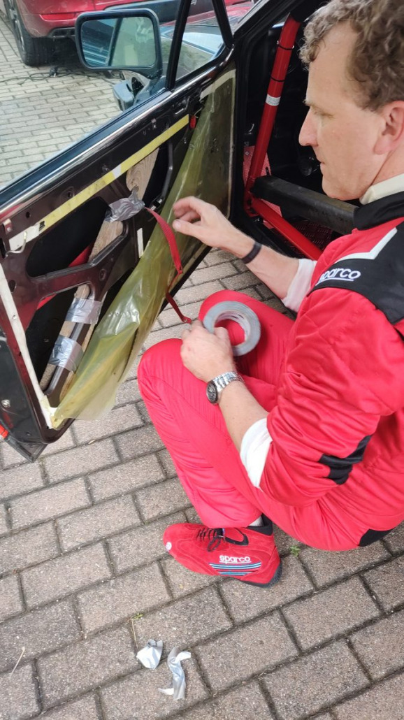 Eine Person in einem roten Rennanzug kniet neben einem Auto, dessen Türinnenverkleidung entfernt wurde. Für die Reparatur verwendet sie rotes Klebeband und durchsichtige Plastikfolie.